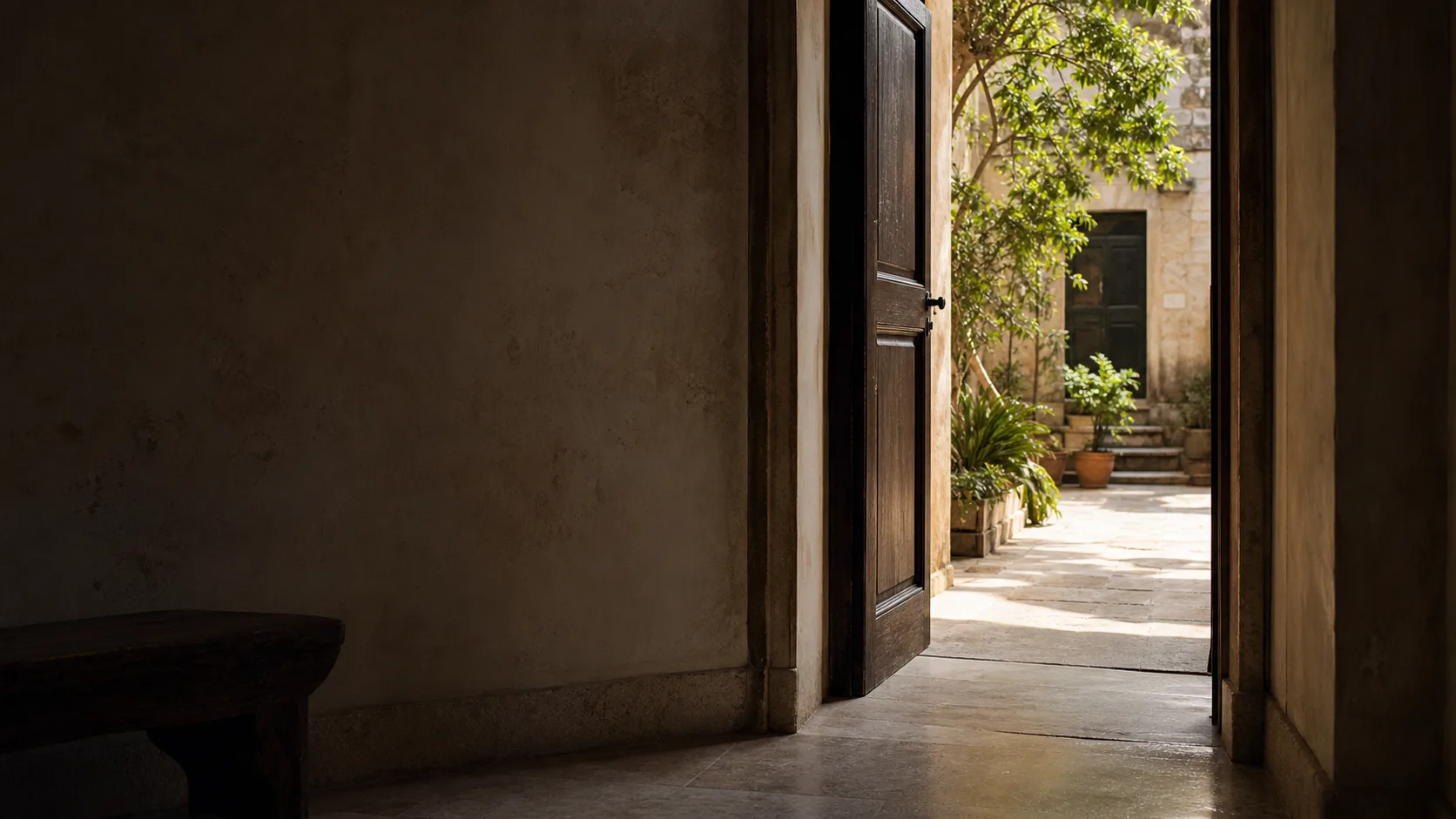 Una porta in legno aperta conduce da un interno in pietra a un cortile illuminato dal sole